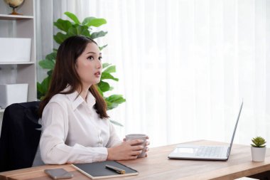 Modern ofis masasındaki Asyalı hevesli genç iş kadını bir fincan kahveyle çalışmak için dizüstü bilgisayar kullanıyor. Çalışkan ve çekici ofis hanımı ofisinde bilgisayar defteri üzerinde çalışıyor..