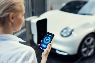 Suit-clad progressive businesswoman look at EV cars battery status from her phone while standing on a charging station with a power cable plug and a renewable energy-powered electric vehicle.