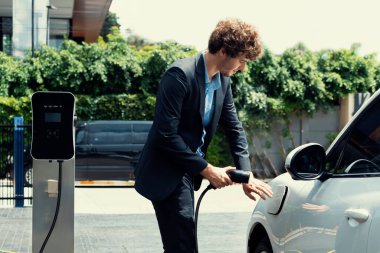 Modern şehir yerleşim bölgesindeki kamu şarj istasyonunda elektrikli araba şarj eden yenilikçi bir işadamı. Çevre dostu şarj edilebilir araba alternatif temiz enerjiyle çalışıyor..