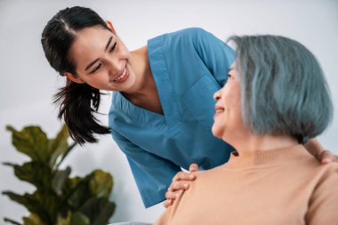 A caregiver rest her hands on the shoulders of a contented senior patient while she sitting on the sofa at home.