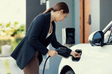 Progressive woman install cable plug to her electric car with home charging station. Concept of the use of electric vehicles in a progressive lifestyle contributes to clean environment.