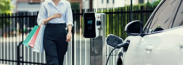 专车在收费站为电动车充电 女商人提着购物袋 城市生活方式的进步与对清洁电能驱动汽车理想的生态关注 — 图库照片