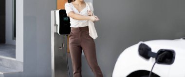 Focus EV charger plugged into EV car at home charging station with blurred background of progressive woman walking in background. Elective vehicle powered by clean energy for eco-friendly concept.