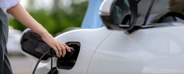 Closeup focus hand insert EV charger to electric vehicle at public charging point in car park with blur business people in backdrop, eco-friendly lifestyle by rechargeable car for progressive concept.
