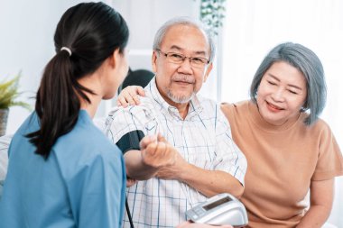 An elderly man having a blood pressure check by his personal caregiver with his wife sitting next to him in their home.
