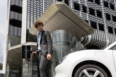 Below view of progressive businessman with electric car recharging at public charging station at modern city residential building. Eco friendly rechargeable EV car powered by alternative clean energy.