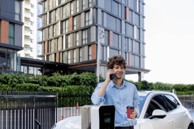 İlerici iş adamı telefonda kahveyle konuşuyor, EV şarj istasyonu ile şarj olan elektrikli arabaya yaslanıyor, yeşil şehir yaşam tarzı olarak arka planda apartman dairesi inşa ediliyor.
