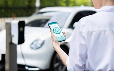 Suit-clad progressive businesswoman look at EV cars battery status from her phone while standing on a charging station with a power cable plug and a renewable energy-powered electric vehicle.