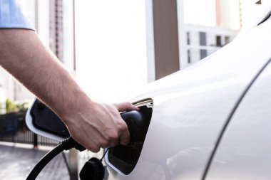 Closeup progressive man holding EV charger plug from public charging station for electric vehicle with background of residential building as concept eco-friendly sustainability energy car concept.