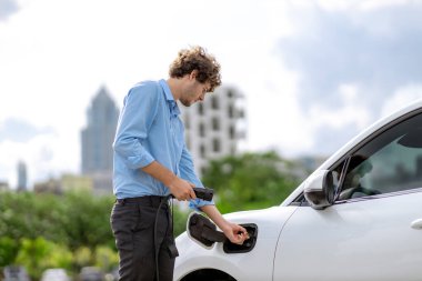 Progressive businessman insert charger plug from charging station to his electric vehicle with apartment condo building in background. Eco friendly rechargeable car powered by sustainable energy.