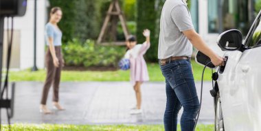 Focus image of progressive man charging electric car from home charging station with blur mother and daughter playing together in the background.