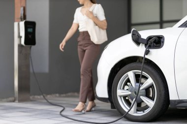 Focus image of electric vehicle recharging battery at home charging station with blurred woman walking in the background. Progressive concept of green energy technology applied in daily lifestyle.