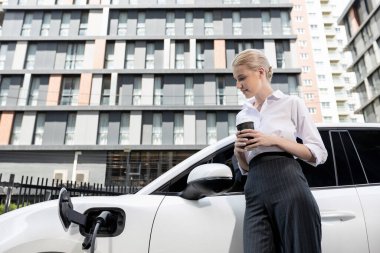 Businesswoman drinking coffee, leaning on electric vehicle recharging at public charging station with residential apartment condos building in background as progressive lifestyle by eco-friendly car.