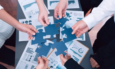 Closeup top view business team of office worker putting jigsaw puzzle together over table filled with financial report paper in workplace with manager to promote harmony concept in meeting room.