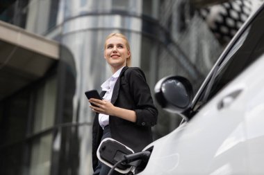 Businesswoman wearing black suit using smartphone, leaning on electric car recharge battery at charging station in city residential building with condos and apartment. Progressive lifestyle concept.