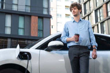 Progressive eco-friendly concept of parking EV car at public electric-powered charging station in city with blur background of businessman leaning on recharging-electric vehicle with coffee.