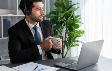 Çağrı merkezi operatörünün müdürü bilgisayarla uğraşırken masasında kahvesiyle oturuyor. Kulaklık takan ve siyah takım giyen bir ofis çalışanı müşteri desteği ya da tele pazarlama üzerinde çalışıyor. coşkulu
