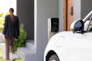 Focus image of electric vehicle recharging battery at home charging station with blurred woman walking in the background. Progressive concept of green energy technology applied in daily lifestyle.