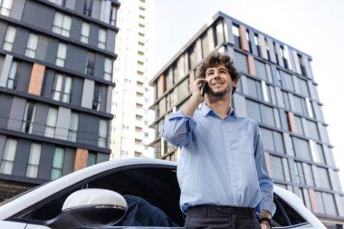 Progressive businessman talking on the phone, leaning on electric car recharging with public EV charging station, apartment condo residential building on the background as green city lifestyle.