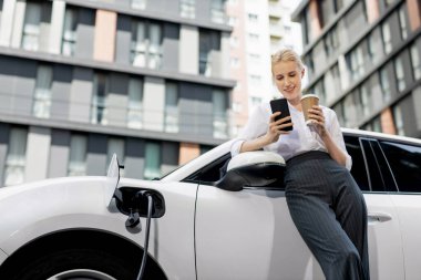 Focus businessman using phone, leaning on electric vehicle, holding coffee with blurred city residential condo buildings in background as progressive lifestyle by renewable and sustainable EV car.
