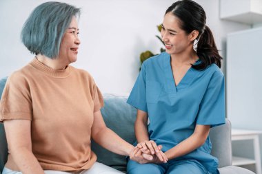 A caregiver rest her hands on the shoulders of a contented senior patient while she sitting on the sofa at home.