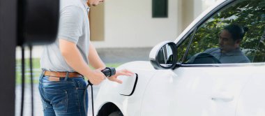 Progressive asian man install cable plug to his electric car with home charging station in the backyard. Concept use of electric vehicles in a progressive lifestyle contributes to clean environment.