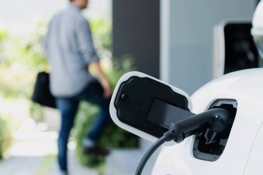 Focus electric car charging at home charging station with blurred progressive man walking in the background. Electric car using renewable clean for eco-friendly concept.