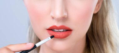 Closeup beautiful young woman with flawless healthy skin and natural makeup putting alluring fashion glossy red lipstick on her lip with lip brush in isolated background.