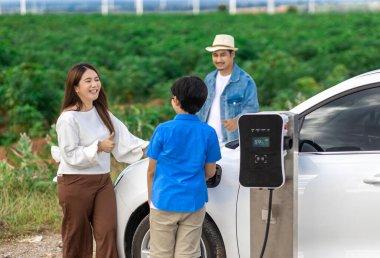 İlerici mutlu aile kavramı elektrikli araçla rüzgar çiftliğinde eğleniyor. Rüzgar türbini jeneratöründen gelen temiz yenilenebilir enerji ile şarj edilen elektrikli araç..