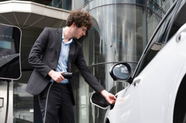 Below view closeup image of progressive black suit businessman recharge battery of his electric vehicle from public charging station. Renewable and alternative energy powered car concept.
