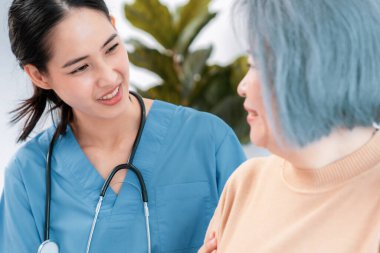 A caregiver rest her hands on the shoulders of a contented senior patient while she sitting on the sofa at home.