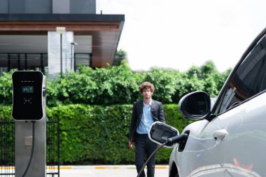 Progressive businessman wearing black suit with electric car recharging at public parking car charging station at modern city residential area. Eco friendly rechargeable car powered by clean energy.