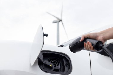 Progressive natural scenic with windmill generator where hand insert charging plug to electric vehicle from charging station with natural background. EV car powered by wind turbine electric generator.