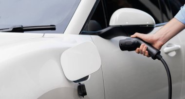 Closeup progressive man holding EV charger plug from public charging station for electric vehicle with background of residential building as concept eco-friendly sustainability energy car concept.