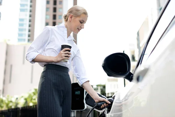 Businesswoman Drinking Coffee Leaning Electric Vehicle Recharging Public Charging Station — Stock Fotó