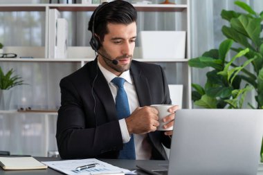Çağrı merkezi operatörünün müdürü bilgisayarla uğraşırken masasında kahvesiyle oturuyor. Kulaklık takan ve siyah takım giyen bir ofis çalışanı müşteri desteği ya da tele pazarlama üzerinde çalışıyor. coşkulu