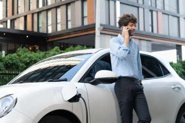 Progressive businessman talking on the phone, leaning on electric car recharging with public EV charging station, apartment condo residential building on the background as green city lifestyle.