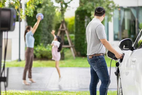 Focus image of progressive man charging electric car from home charging station with blur mother and daughter playing together in the background.