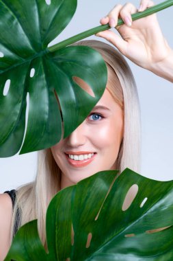 Closeup facial portrait personable woman with perfect smooth makeup holding green monstera leaves and cover her face as natural healthy skincare treatment. Tropical nature and beauty concept