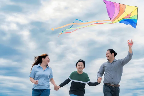 Progressive happy family vacation and carefree day concept. Young parents mother father and son run along and flying kite together road with enjoy natural scenic on scenery and clear sky background.