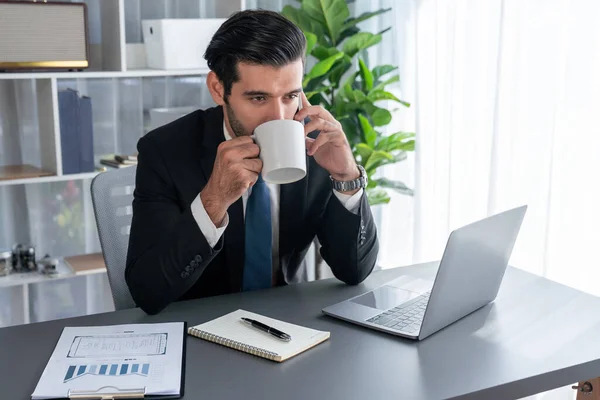 Businessman working in modern office workspace with cup of coffee in his hand while answering phone call making sales calls or managing employee. Fervent