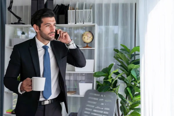 Diligent and busy businessman talking on phone call with coworker in modern office. Making sales call to client with coffee in hand, standing posing confidently as professional office worker. Fervent