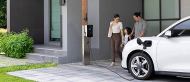 Progressive young parents and daughter with electric vehicle and home charging station. Green and clean energy from electric vehicles for healthy environment. Eco power from renewable source at home.