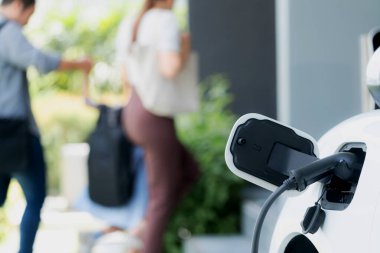 Focus closeup electric vehicle recharging battery from home electric charging station with blurred family in background. Renewable clean energy car for progressive eco awareness lifestyle concept.