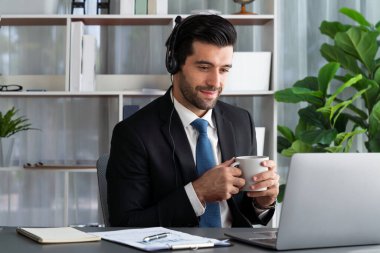 Çağrı merkezi operatörünün müdürü bilgisayarla uğraşırken masasında kahvesiyle oturuyor. Kulaklık takan ve siyah takım giyen bir ofis çalışanı müşteri desteği ya da tele pazarlama üzerinde çalışıyor. coşkulu