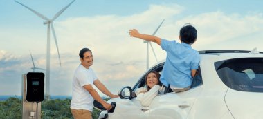 İlerici mutlu aile kavramı elektrikli araçla rüzgar çiftliğinde eğleniyor. Rüzgar türbini jeneratöründen gelen temiz yenilenebilir enerji ile şarj edilen elektrikli araç..