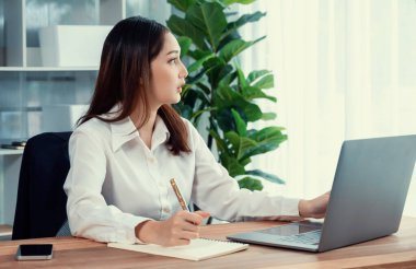 Modern ofis masasındaki Asyalı hevesli genç iş kadını çalışmak ve notlar yazmak için dizüstü bilgisayar kullanıyor. Çalışkan ve çekici ofis hanımı ofisinde bilgisayar defteri üzerinde çalışıyor..