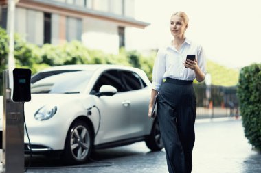 İş kadını tablet kullanıyor, halka açık otoparkta elektrikli aracını şarj ederken yürüyor. Teknolojinin ilerici yaşam tarzı ve EV otomobilinin ekolojik kaygısı