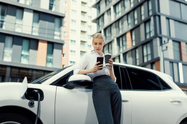 Focus businessman using phone, leaning on electric vehicle, holding coffee with blurred city residential condo buildings in background as progressive lifestyle by renewable and sustainable EV car.