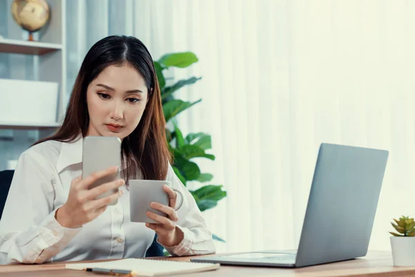 現代的で若い熱心なビジネス女性のマルチタスクは ビジネス上の問題のために彼女のスマートフォンをチェックし オフィスで彼女の机の上でノートパソコンで作業することによって — ストック写真